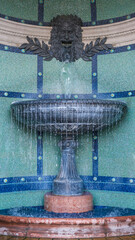 Decorative stone fountain with a lion-faced spout pouring water into a tiled basin, set against a mosaic wall in Budapest.