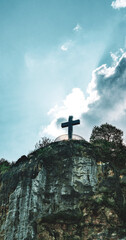 Large metal cross standing atop a rocky hill with dramatic backlighting from the sun behind clouds,...