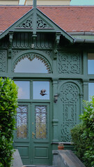 Ornate green wooden door and facade with decorative carvings and wrought-iron details, partially framed by green bushes.