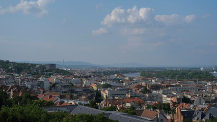 Panoramic view of Budapest rooftops and the Danube River stretching toward the horizon, under a lightly clouded sky.
