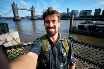 Happy male tourist with backpack taking a selfie with tower bridge in the background, enjoying summer holidays in london, united kingdom, with city hall visible on the right