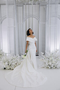 Bride in a flowing white gown holding a flower while posing on an ornate all-white wedding stage with floral decorations. Elegant Bride in White Gown Posing on Luxurious Wedding Stage
