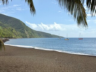 view of a black sand beach in Guadeloupe in the French Antilles with sailboats on the Caribbean Sea and a mountain overlooking the sea
