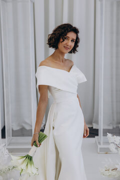 Bride in a flowing white gown holding a flower while posing on an ornate all-white wedding stage with floral decorations. Elegant Bride in White Gown Posing on Luxurious Wedding Stage