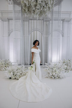 Bride in a flowing white gown holding a flower while posing on an ornate all-white wedding stage with floral decorations. Elegant Bride in White Gown Posing on Luxurious Wedding Stage