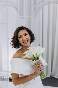 Bride in a flowing white gown holding a flower while posing on an ornate all-white wedding stage with floral decorations. Elegant Bride in White Gown Posing on Luxurious Wedding Stage