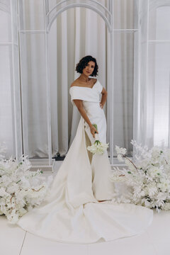 Bride in a flowing white gown holding a flower while posing on an ornate all-white wedding stage with floral decorations. Elegant Bride in White Gown Posing on Luxurious Wedding Stage