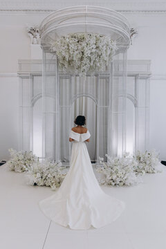 Bride in a flowing white gown holding a flower while posing on an ornate all-white wedding stage with floral decorations. Elegant Bride in White Gown Posing on Luxurious Wedding Stage