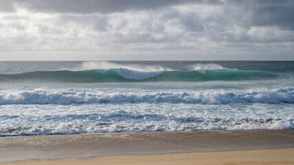 Powerful ocean wave rolls toward shore under dramatic clouds, captured from beach level with visible spray.