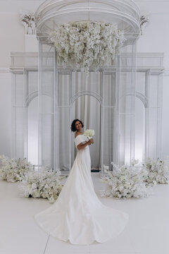 Bride in a flowing white gown holding a flower while posing on an ornate all-white wedding stage with floral decorations. Elegant Bride in White Gown Posing on Luxurious Wedding Stage