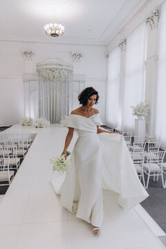 Bride in a flowing white gown holding a flower while posing on an ornate all-white wedding stage with floral decorations. Elegant Bride in White Gown Posing on Luxurious Wedding Stage