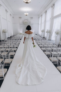 Bride in a flowing white gown holding a flower while posing on an ornate all-white wedding stage with floral decorations. Elegant Bride in White Gown Posing on Luxurious Wedding Stage