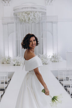 Bride in a flowing white gown holding a flower while posing on an ornate all-white wedding stage with floral decorations. Elegant Bride in White Gown Posing on Luxurious Wedding Stage