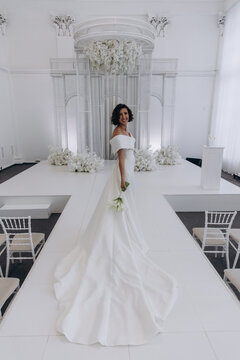 Bride in a flowing white gown holding a flower while posing on an ornate all-white wedding stage with floral decorations. Elegant Bride in White Gown Posing on Luxurious Wedding Stage
