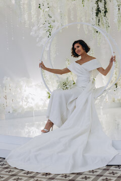 Elegant bride in a satin gown posing on a circular swing surrounded by cascading white flowers and luxurious wedding decor. Glamorous Bride Posing on Decorative Swing with Floral Backdrop