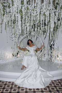 Elegant bride in a satin gown posing on a circular swing surrounded by cascading white flowers and luxurious wedding decor. Glamorous Bride Posing on Decorative Swing with Floral Backdrop