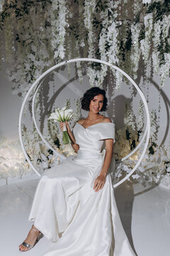 Elegant bride in a satin gown posing on a circular swing surrounded by cascading white flowers and luxurious wedding decor. Glamorous Bride Posing on Decorative Swing with Floral Backdrop