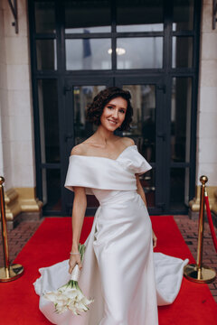 Glamorous bride in an off-shoulder wedding dress holding a calla lily bouquet, posing gracefully on a red carpet in front of a grand building. Elegant Bride on Red Carpet Holding Calla Lily Bouquet