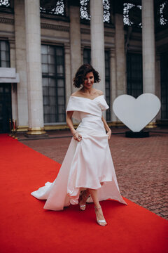 Glamorous bride in an off-shoulder wedding dress holding a calla lily bouquet, posing gracefully on a red carpet in front of a grand building. Elegant Bride on Red Carpet Holding Calla Lily Bouquet