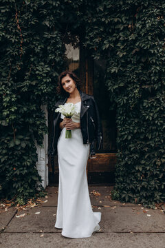 Full-length portrait of a bride in a white gown and leather jacket holding flowers while standing near a lush ivy-covered wall. Bride in Leather Jacket Holding Flowers Near Ivy Wall