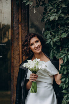 Full-length portrait of a bride in a white gown and leather jacket holding flowers while standing near a lush ivy-covered wall. Bride in Leather Jacket Holding Flowers Near Ivy Wall