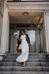 Stylish bride wearing a white wedding dress and a black leather jacket posing confidently on stone...