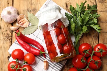 Tasty pickled tomatoes in jar and ingredients on wooden table, flat lay