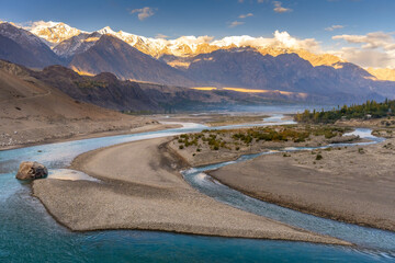 A turquoise river winds through a rocky valley, framed by snow-capped mountains glowing in golden...