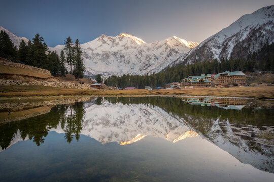 Golden sunlight touches snowy peaks reflected in a calm alpine lake, with wooden lodges nestled among pine trees, creating a peaceful mountain retreat scene under a clear sky