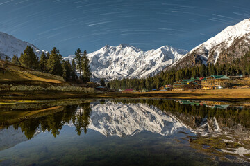 A stunning night view of Nanga Parbat’s snow-covered peak reflected in a still alpine lake, with surrounding pine trees and star trails illuminating the clear night sky in Fairy Meadows.