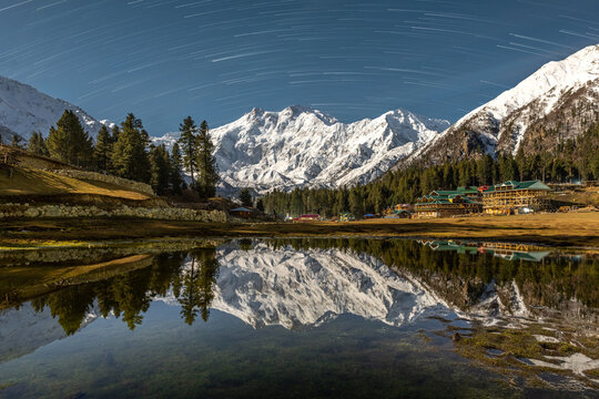 A stunning night view of Nanga Parbat&rsquo;s snow-covered peak reflected in a still alpine lake, with surrounding pine trees and star trails illuminating the clear night sky in Fairy Meadows.