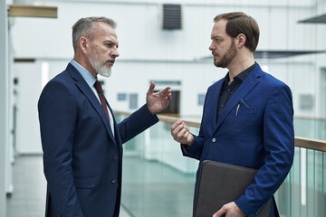 Two businessmen engaging in serious conversation in contemporary office setting, one holding document folder. Professional attire and modern interior create formal atmosphere