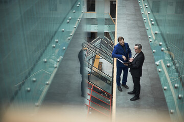 Obraz premium Two businessmen reading and discussing document in a modern office environment with a glass railing and multiple levels, highlighting professional attire and conversational engagement