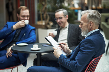Fototapeta premium Three businessmen sitting at a round table discussing plans during a coffee break in a modern office setting. All holding cups of coffee while engaged in conversation