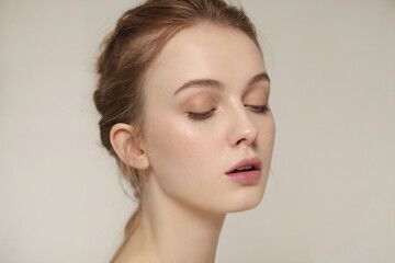 A soft, minimalist beauty portrait against a plain off-white background. The subject has fair skin, closed eyes with natural-looking nude eyeshadow, and a gentle pink lip color.