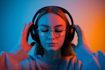 A close-up portrait shot of a young woman wearing large black over-ear headphones and round glasses, with closed eyes in a peaceful listening pose. 