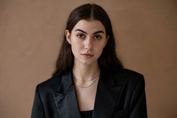 A professional headshot portrait against a plain beige background. The subject has long dark brown hair parted in the middle, wearing a black blazer over a black top.