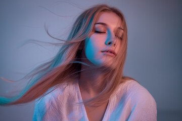 A moody portrait photograph with blue and pink lighting. A young woman has long blonde hair flowing dramatically to the left side of the frame, caught in motion.