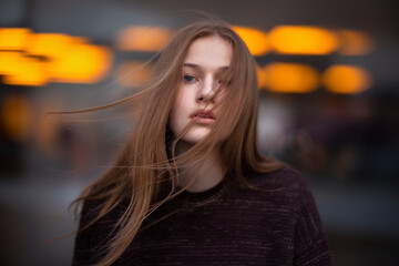 A moody portrait photograph of a young woman with long straight brown hair blowing in the wind against a blurred background of orange bokeh lights. 