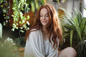 A soft, natural portrait photograph of a young woman with long, wavy auburn hair in a bright indoor setting with plants.
