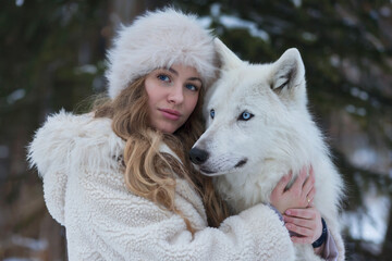 A winter portrait photograph of a young woman in a white furry ushanka hat and a cream-colored knit sweater, embracing a white arctic wolf.