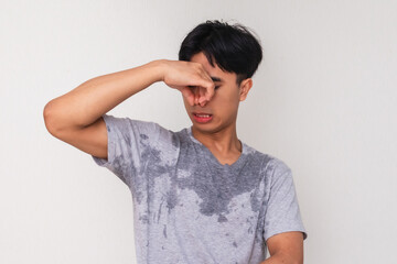Young Asian man sweating and closing his nose with a disgusted face due to body odor, isolated on white background, perfect for hygiene or deodorant advertisement