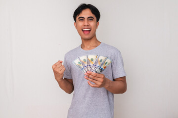 Portrait of a happy young Asian man holding cash and clenching his fist in celebration, smiling...