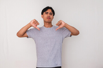 Angry young Asian man showing thumbs down gesture with mad expression, standing against white...