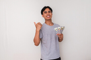 Happy young Asian man holding money and pointing thumb behind him with cheerful expression,...