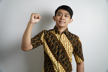 A smiling Asian man in a traditional batik shirt and black peci flexes his bicep, showcasing...