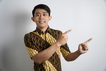 smiling Asian man in a traditional batik shirt stands confidently, pointing sideways with both...