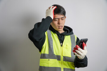 A frustrated Asian man in a high-visibility safety vest and work gloves holds his phone while...