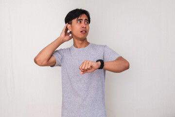 Portrait of a confused young Asian man looking at his wristwatch while standing against a white...