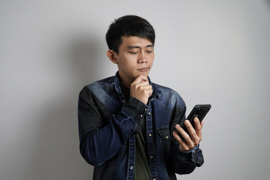 A thoughtful young Asian man holds a smartphone with a hand on his chin